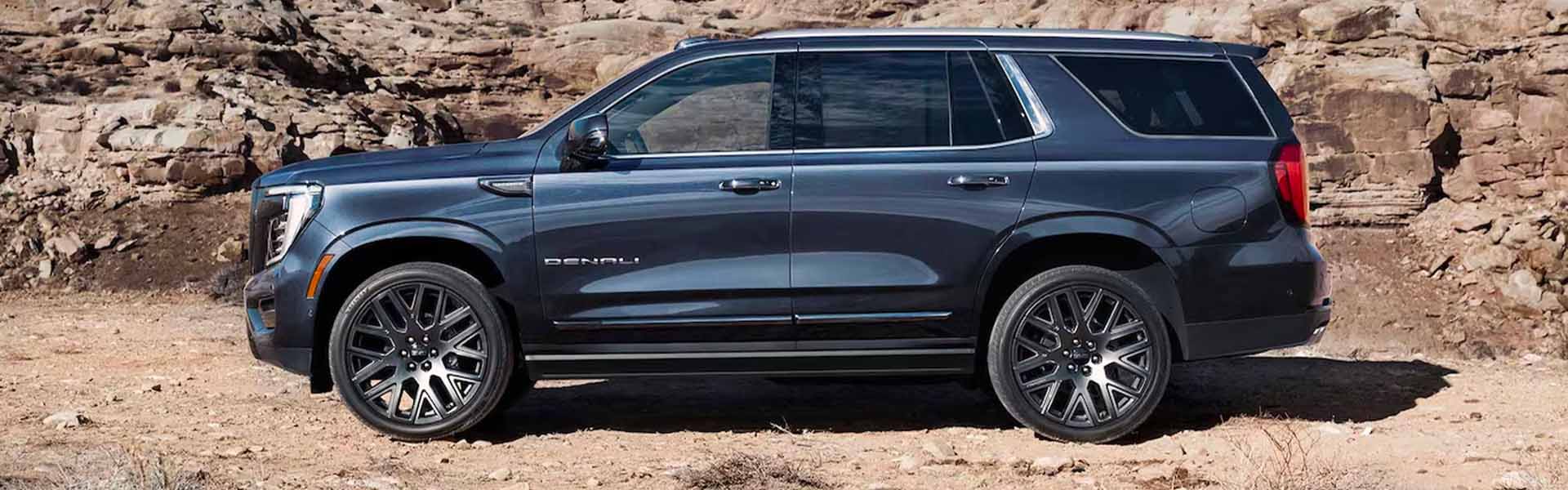 2025 GMC Yukon full-size SUV in dark blue shown in side profile on a rocky desert trail with rugged terrain in the background.