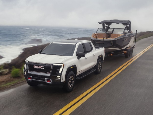 **Alt text:** White 2026 GMC Sierra EV towing a boat on a trailer along a coastal highway, with ocean waves and foggy skies in the background.