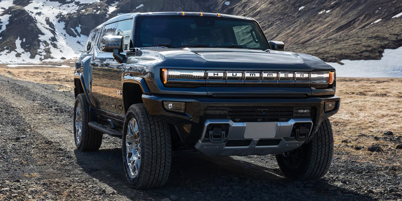 A black 2026 GMC Hummer EV SUV truck is parked on a rocky path in a mountainous landscape with patches of snow. The sky is overcast, creating a rugged, adventurous mood.